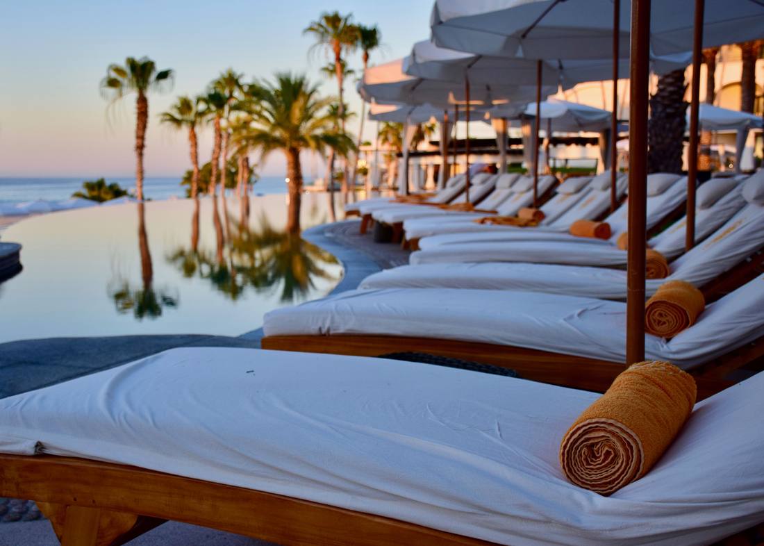 White sun loungers beside an infinity pool at sunset, palms reflected in the water