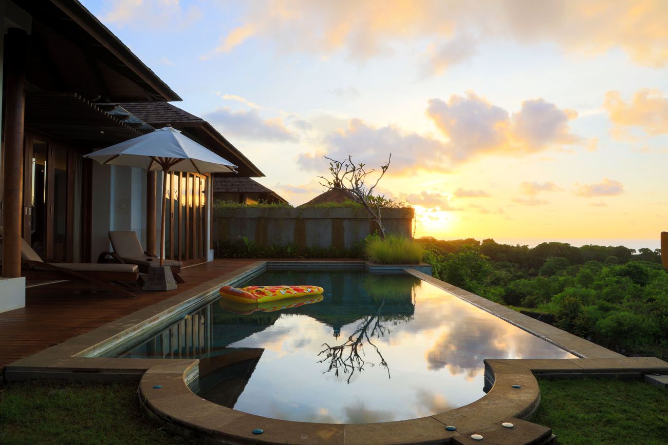 A pool at sunset reflecting golden clouds, with loungers under an umbrella