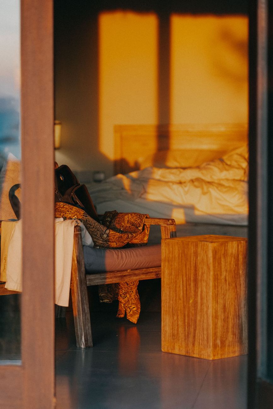 Late sun pouring into a bedroom across a teak bench and an unmade bed