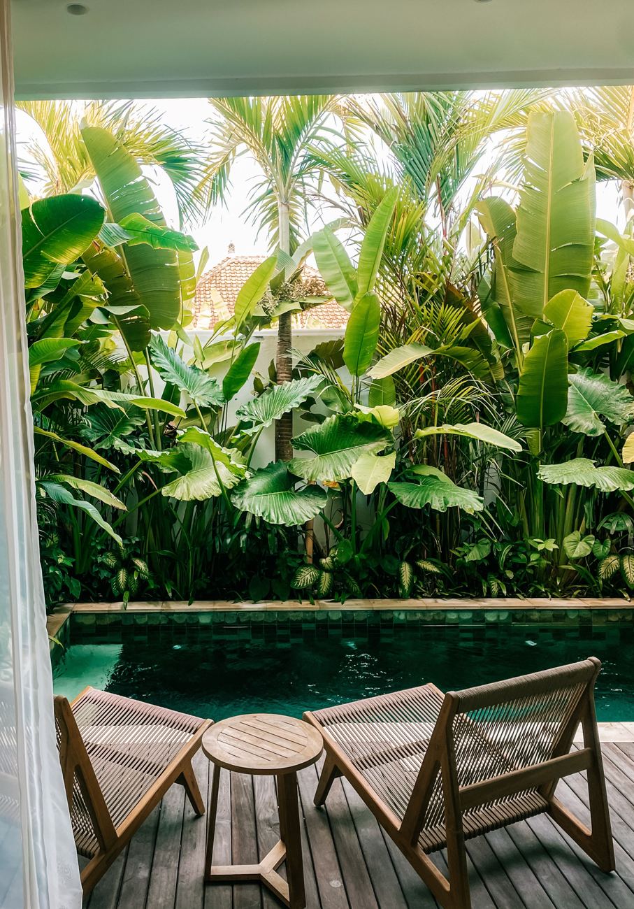 Two woven teak chairs on a wooden deck beside a green-tiled pool, framed by banana leaves and palms